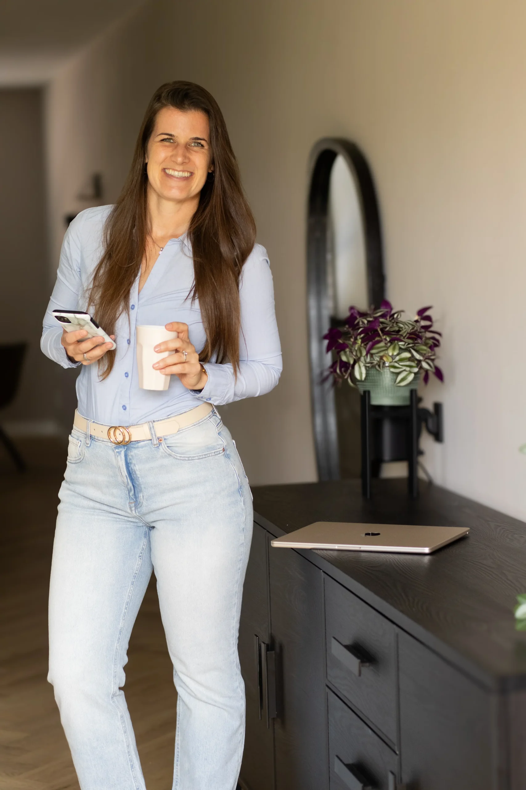 VA Britt lachend met telefoon en beker, laptop en plant op tafel.